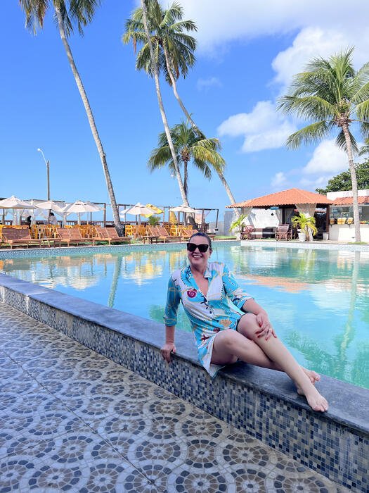 Mulher à beira da piscina de uma pousada, cercada de coqueiros